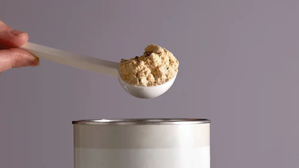 Close-up of a hand holding a plastic scoop filled with protein powder over an open metal can.